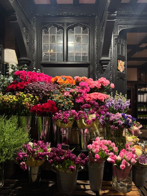 A display of fresh flowers in various vibrant colors, including pink, orange, red, purple, and yellow, arranged in metallic and glass vases on a dark wooden surface inside a florist shop. The flowers