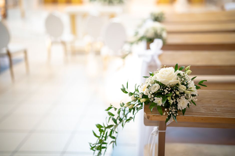 A close-up of a white floral bouquet resting on a wooden pew in an indoor wedding setting, featuring a large white rose, smaller white flowers, green foliage, and delicate baby's breath, arranged with