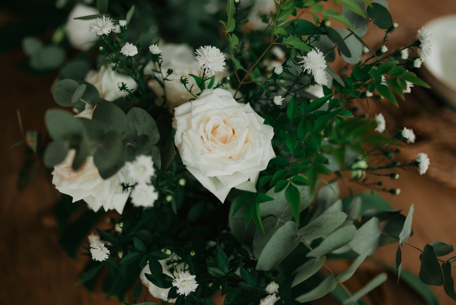 A floral arrangement featuring a central ivory rose surrounded by sprigs of small white daisies and delicate white baby's breath, all set within an assortment of lush green foliage including eucalyptu
